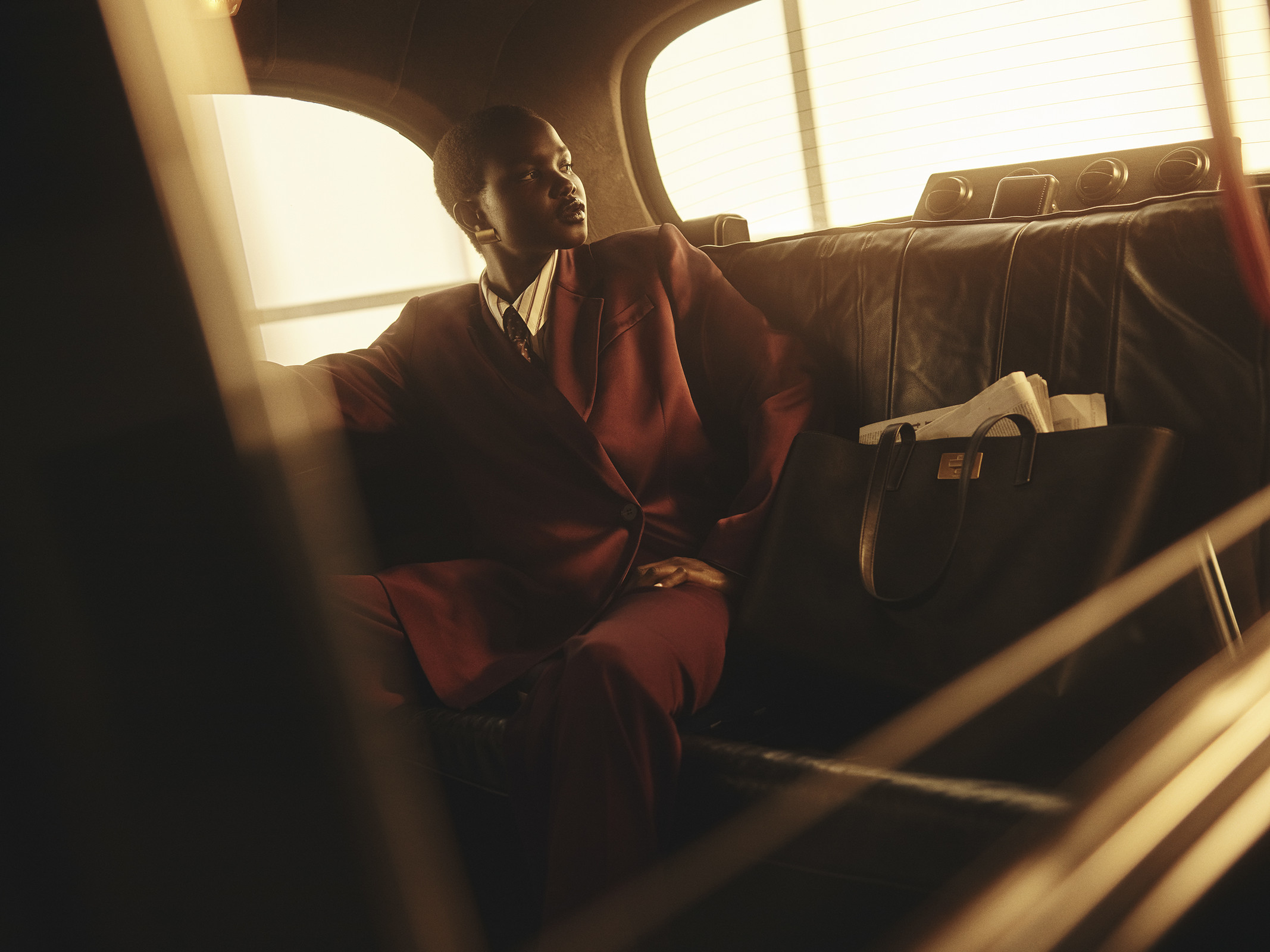 A woman in a maroon suit sits in a taxi with a black leather bag and a newspaper, illuminated by warm light from a window.