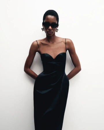 Portrait of an African American woman posing in a formal black dress, sunglasses, and dangle earrings in front of a plain white wall.