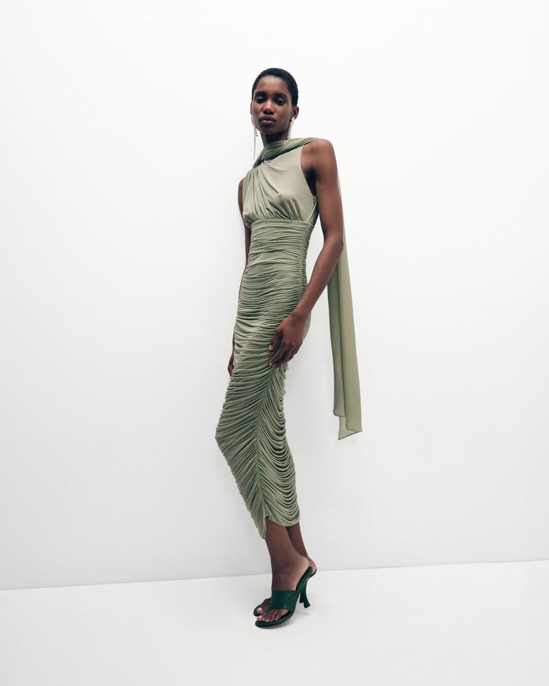Elegant woman in a draped olive green dress and matching heeled mules strikes a pose against a stark white background, highlighting the dress's texture.