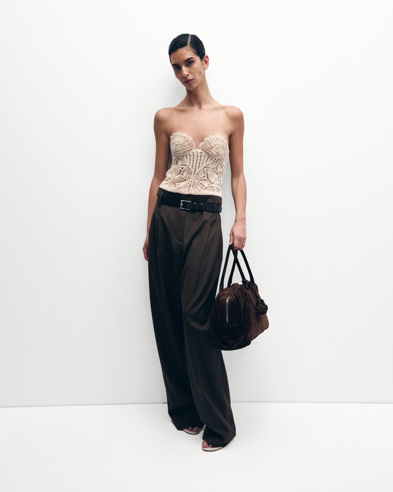 Fashion shot of a woman wearing a tan bustier top and brown pants, holding a matching handbag, against a white backdrop.