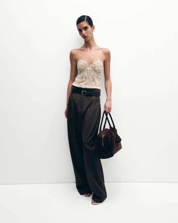 Fashion shot of a woman wearing a tan bustier top and brown pants, holding a matching handbag, against a white backdrop.