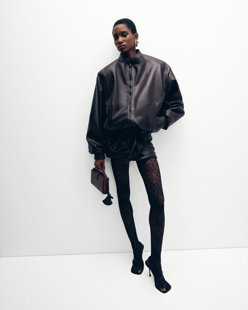 An African-American woman stands confidently in a studio, wearing a leather jacket, shorts, patterned tights, and heels, holding a purse.