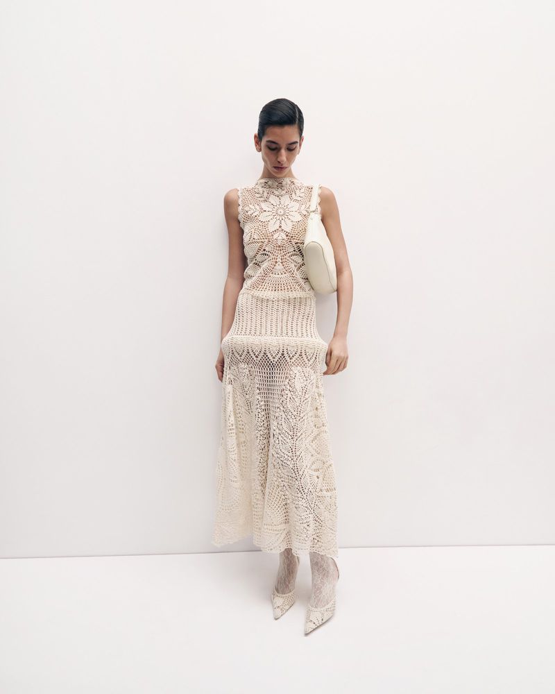 A slender woman poses against a white wall, showcasing a cream-colored, sleeveless lace dress with matching heels and a white shoulder bag.