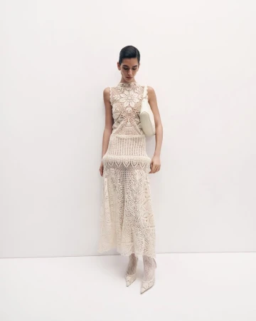 A slender woman poses against a white wall, showcasing a cream-colored, sleeveless lace dress with matching heels and a white shoulder bag.