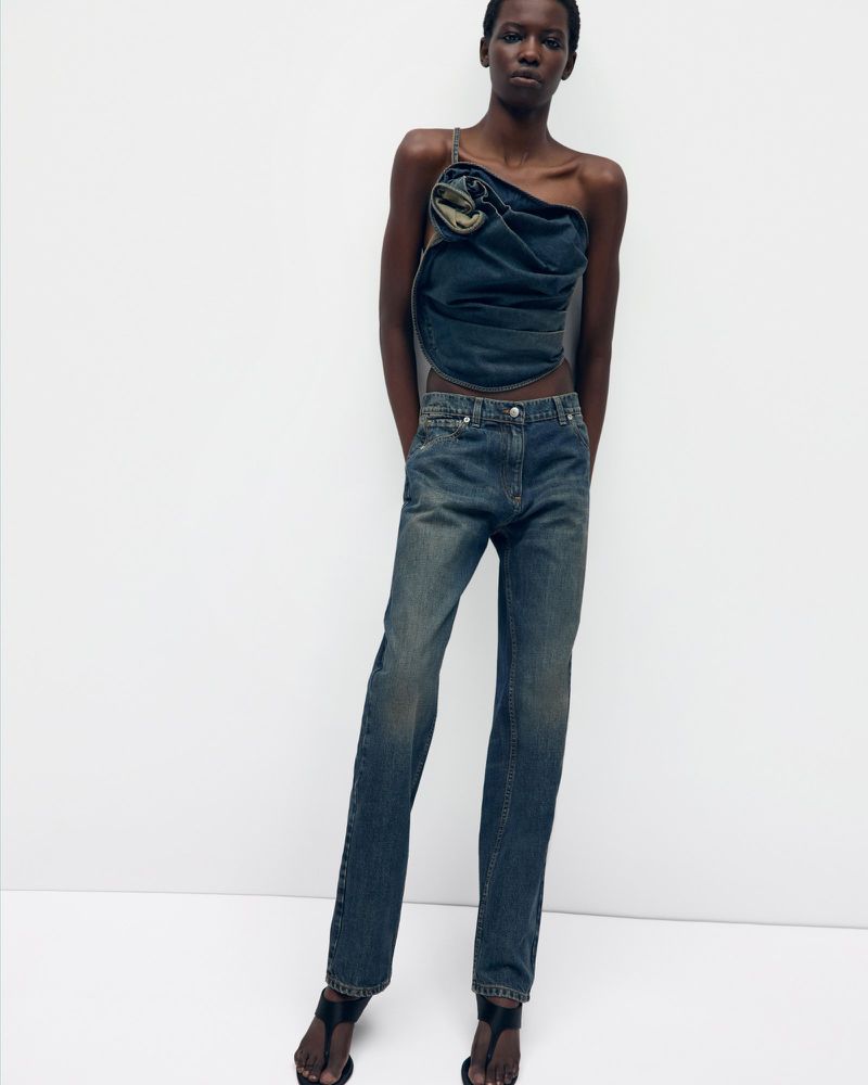 A fashion shot of an adult woman posing in a blue denim top and jeans in front of a white wall in a photo studio.