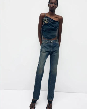 A fashion shot of an adult woman posing in a blue denim top and jeans in front of a white wall in a photo studio.