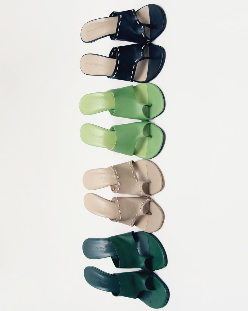 Arrangement of four pairs of leather toe-ring sandals in shades of dark blue, green, beige, and dark green on a white backdrop.