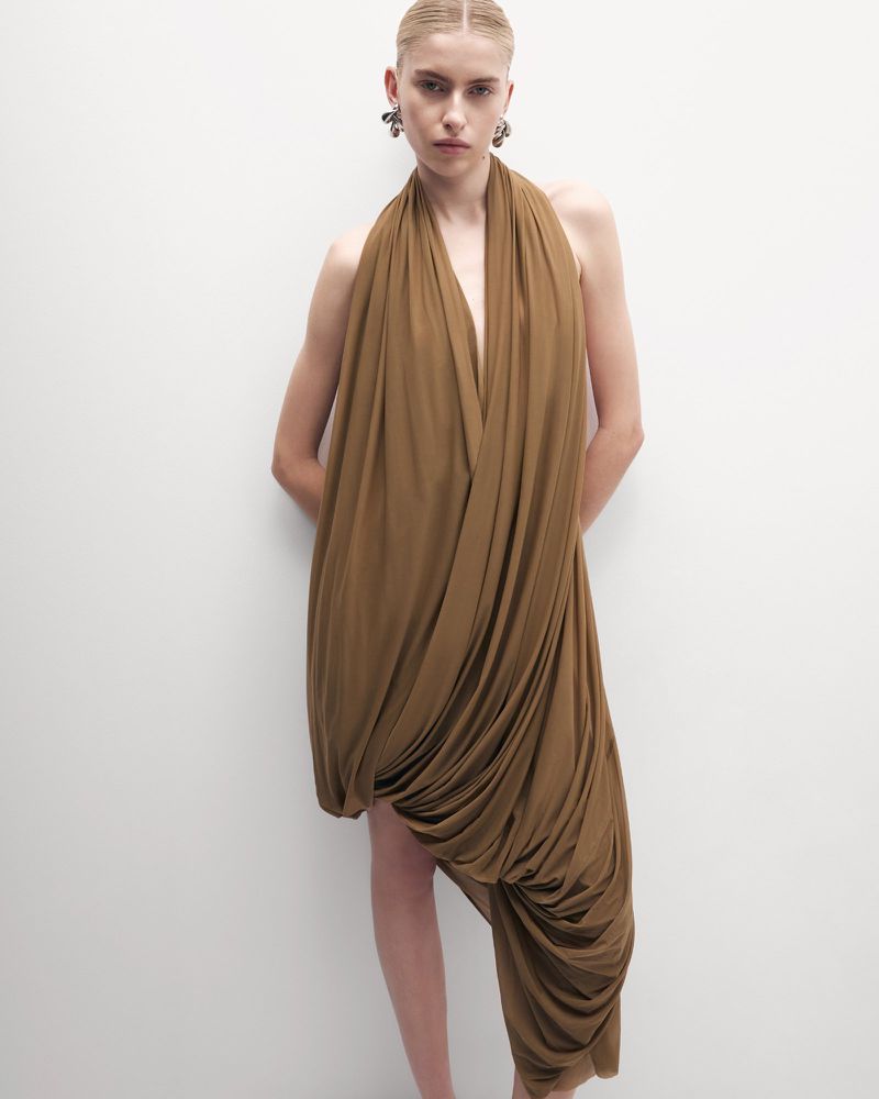 Woman wearing a draped, halter-style dress in a studio setting, minimalist fashion, stylish earrings, and neutral background.
