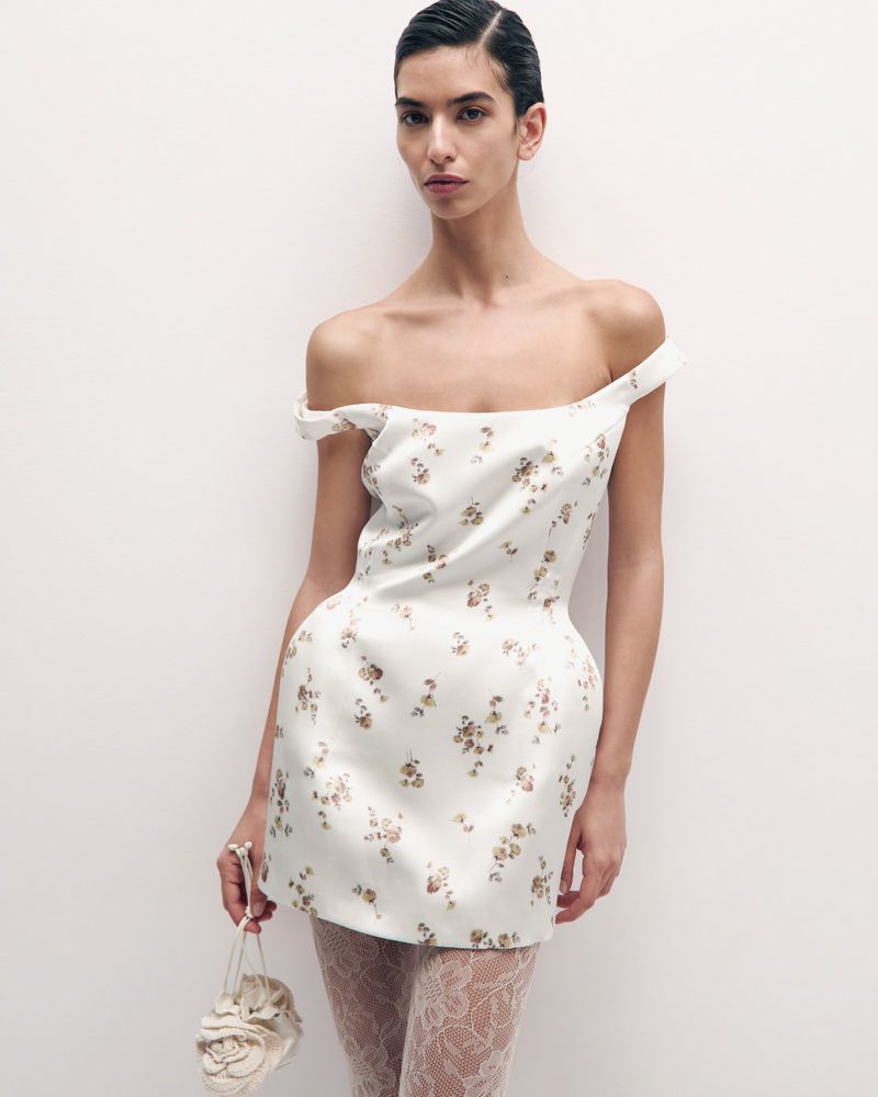 A female fashion model in an off-the-shoulder white floral dress with white lace tights, holding a flower purse against a plain white backdrop.