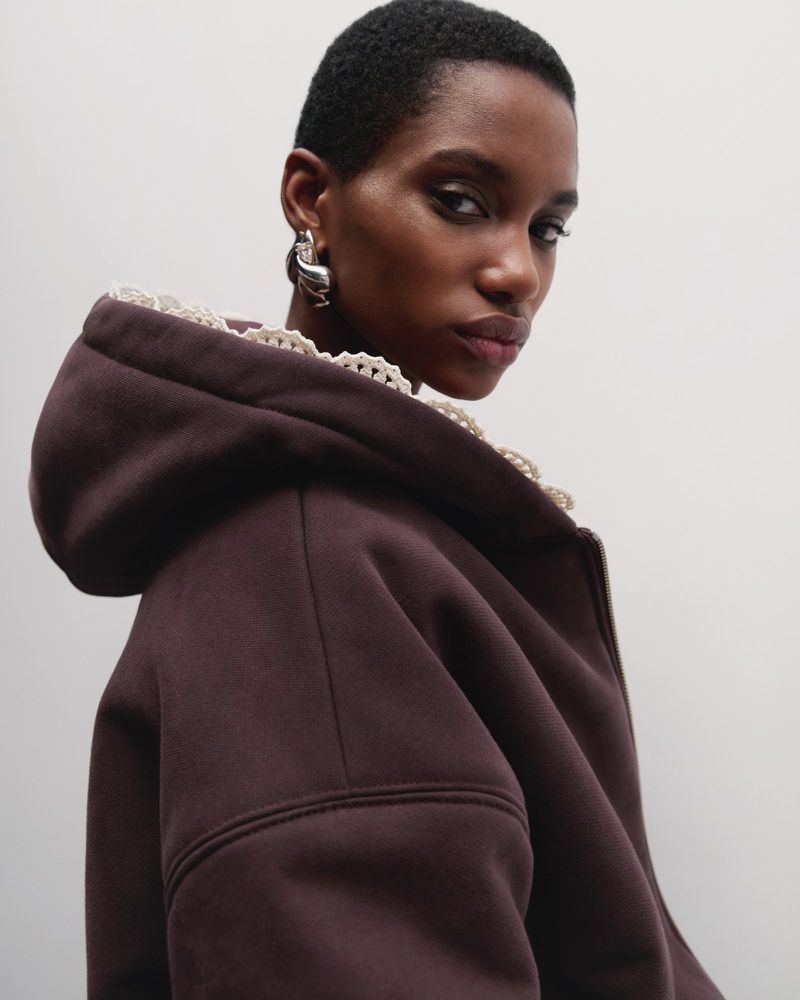 Close-up of a stylish African-American woman with short hair wearing a brown hooded top and silver earrings against a white backdrop.