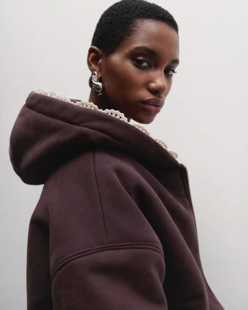 Close-up of a stylish African-American woman with short hair wearing a brown hooded top and silver earrings against a white backdrop.