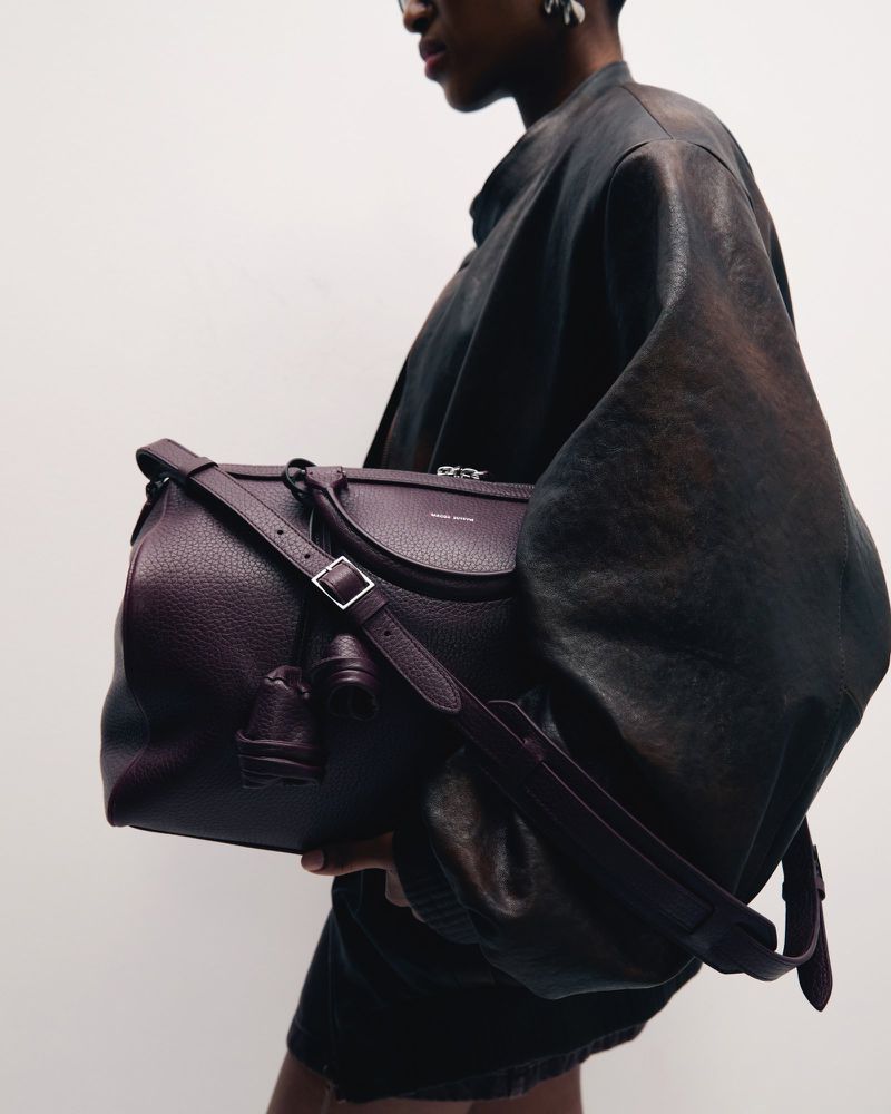 Fashion shot of a dark-skinned adult wearing a dark brown jacket and holding a large maroon leather handbag with a strap against a white background.