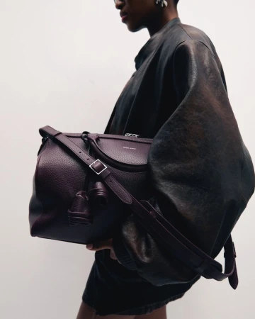 Fashion shot of a dark-skinned adult wearing a dark brown jacket and holding a large maroon leather handbag with a strap against a white background.