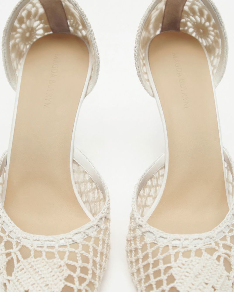 Close-up of two beige high-heeled shoes with crocheted vamps set against a bright white background, showcasing the delicate lace design.