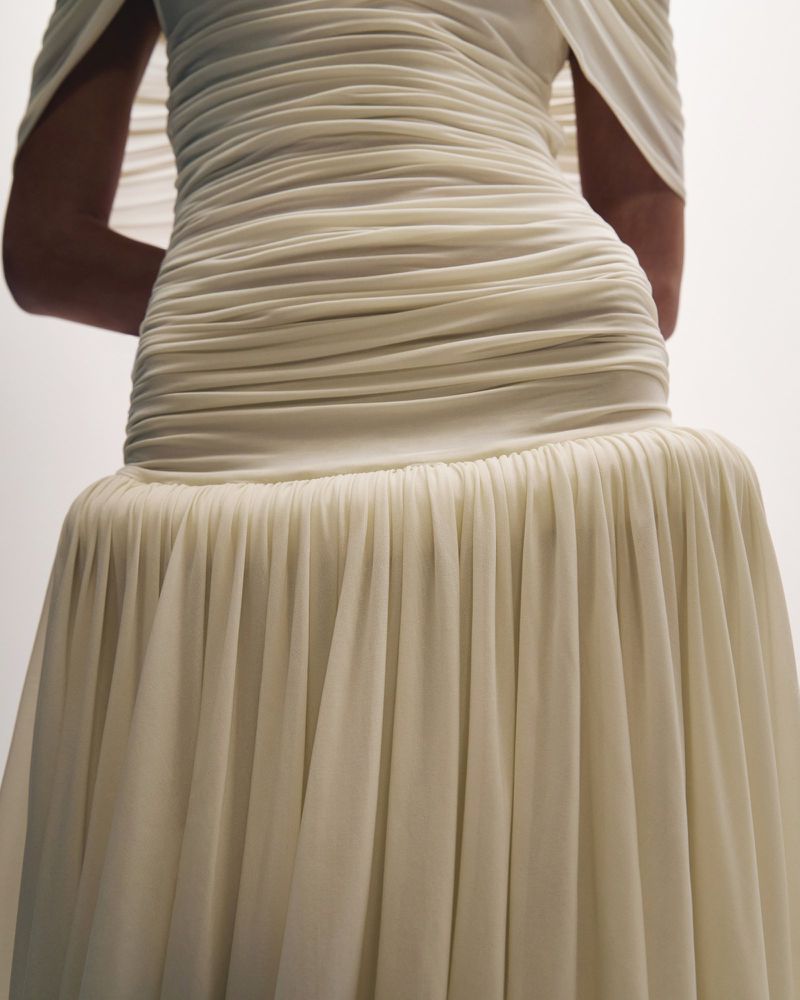 Close-up of an adult female wearing an elegant, pleated, off-white couture gown with detailed draping and flowing skirt against a plain background.