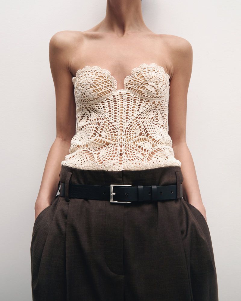 Woman wearing a cream crochet top and dark brown, wide-legged pants with a black belt, posing in a studio against a white background.
