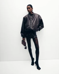 Model in a dark leather jacket and patterned tights holding a small handbag against a plain white studio backdrop