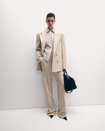 Model standing against a white seamless background wearing a beige oversized suit jacket and holding a dark handbag.