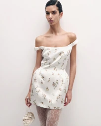 Model in an off-the-shoulder floral mini dress and lace tights holding a small handbag against a plain white background.