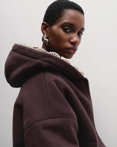 Close-up of a model wearing a dark brown hooded jacket with lace trim and a silver sculptural earring against a grey studio backdrop.