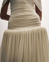 Close-up of a model wearing a cream ruched drop-waist gown with a gathered pleated skirt against a white studio background.