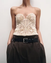 Model wearing a cream crochet strapless top and dark brown trousers with a black leather belt against a neutral white backdrop.
