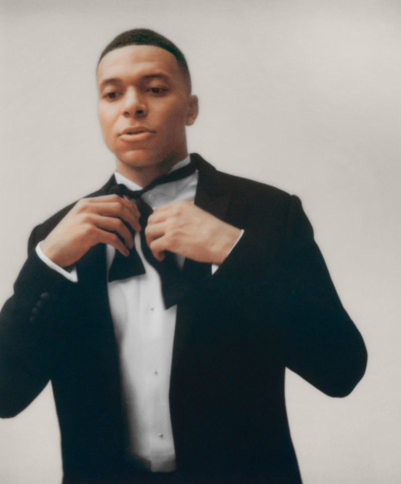 Portrait of a man in a tuxedo adjusting a bow tie against a neutral backdrop, exuding formal elegance in a studio-like setting.