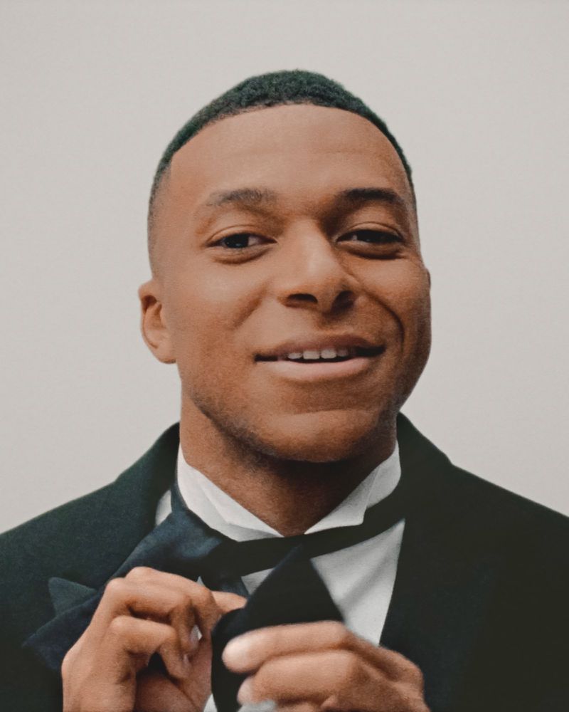 Close-up portrait of a smiling adult male wearing a black tuxedo and adjusting a black bow tie against a plain light background.