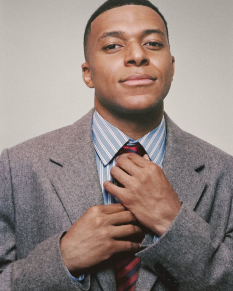Portrait of a young adult male in a grey blazer and striped shirt, adjusting a red and blue striped tie, against an off-white background.