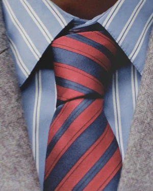 Close up of a red and navy striped tie paired with a light blue striped collared shirt and grey textured jacket lapels.