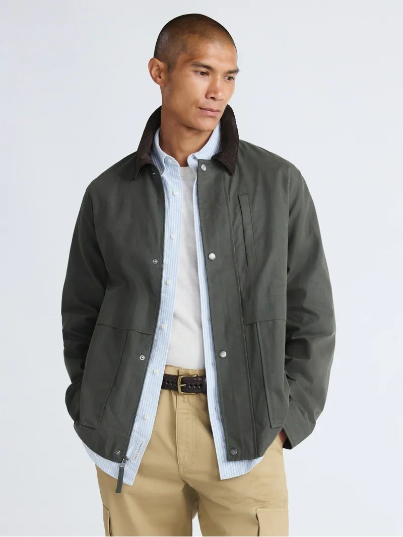 Portrait of an adult male wearing a green jacket, light blue shirt, and khaki pants in a studio setting with soft, diffused lighting.