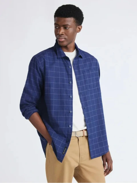 A dark-skinned adult male is posing indoors, wearing an open blue and white checkered shirt over a white t-shirt and khaki pants with a tan belt.