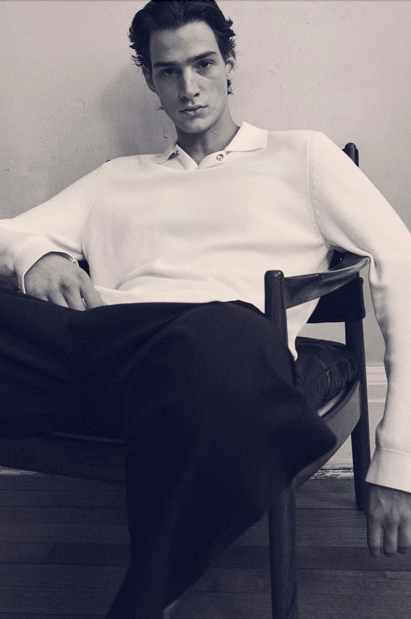 Noah Herbst seated in a wooden armchair wearing a white long-sleeve knit polo shirt and black trousers for COS.
