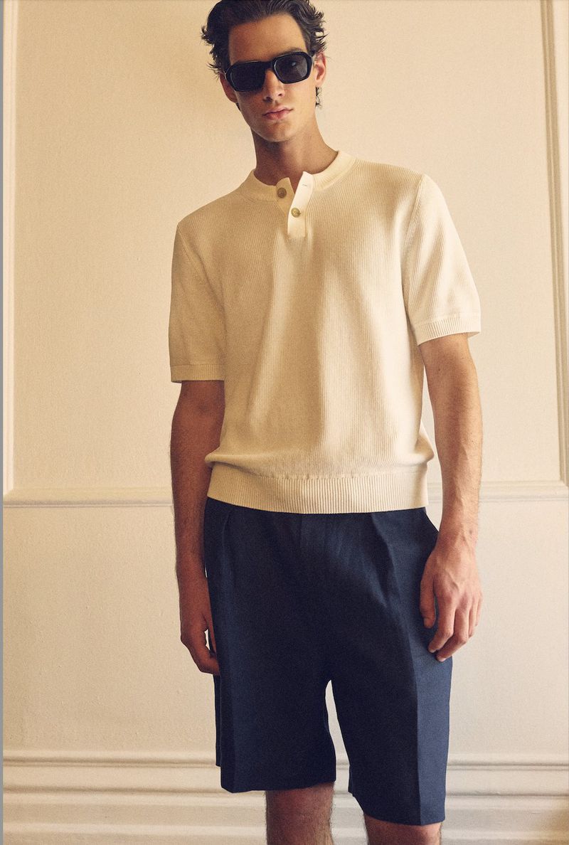 A fair-skinned man poses in front of a neutral wall, sporting sunglasses, a cream knit shirt, and navy pleated shorts for a casual style.