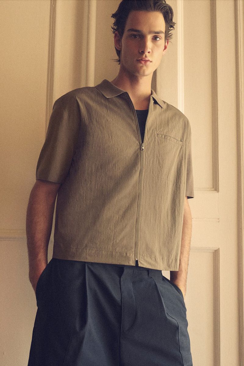 A young adult man poses indoors in a short-sleeved khaki shirt with a zipper and dark blue dress pants, standing against a cream door.