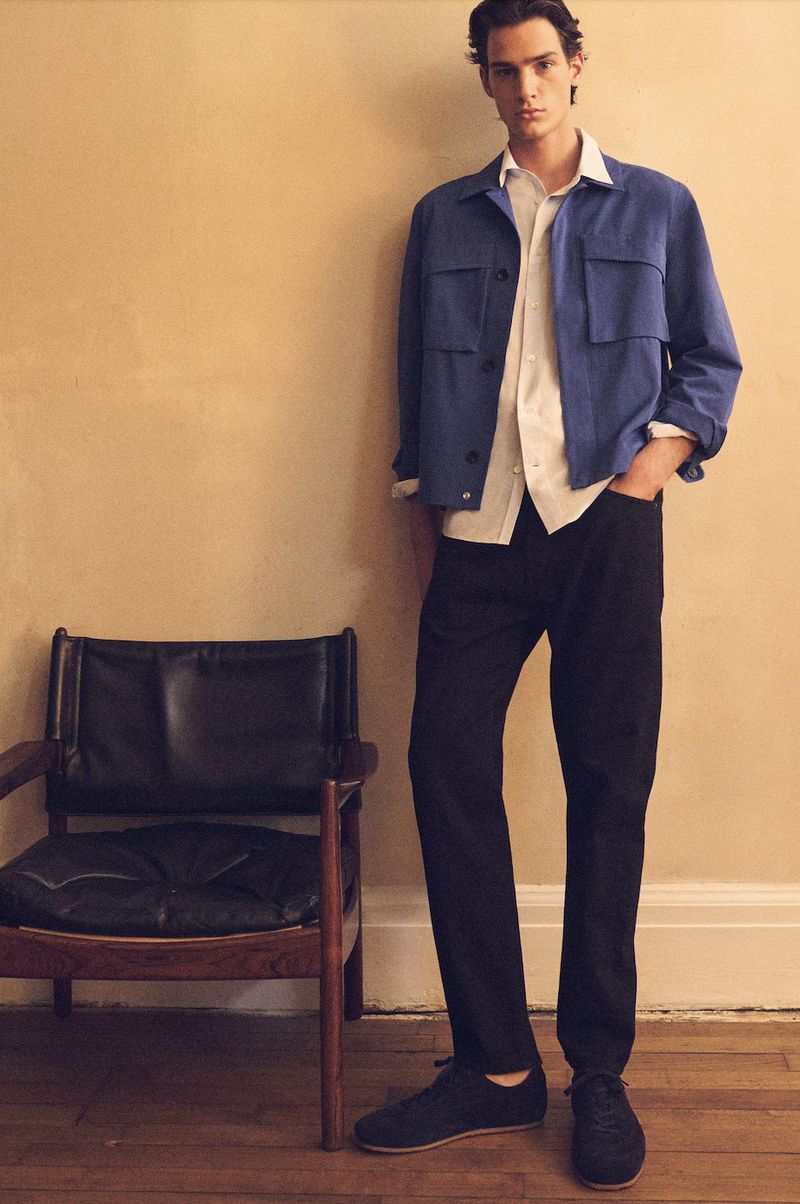 Male fashion model in a blue jacket and black pants posing indoors in a neutral setting with a wood floor and mid-century chair.