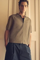 Model Noah Herbst in a beige zip-up short-sleeve shirt and dark navy trousers for the COS Pre Fall 2025 campaign.
