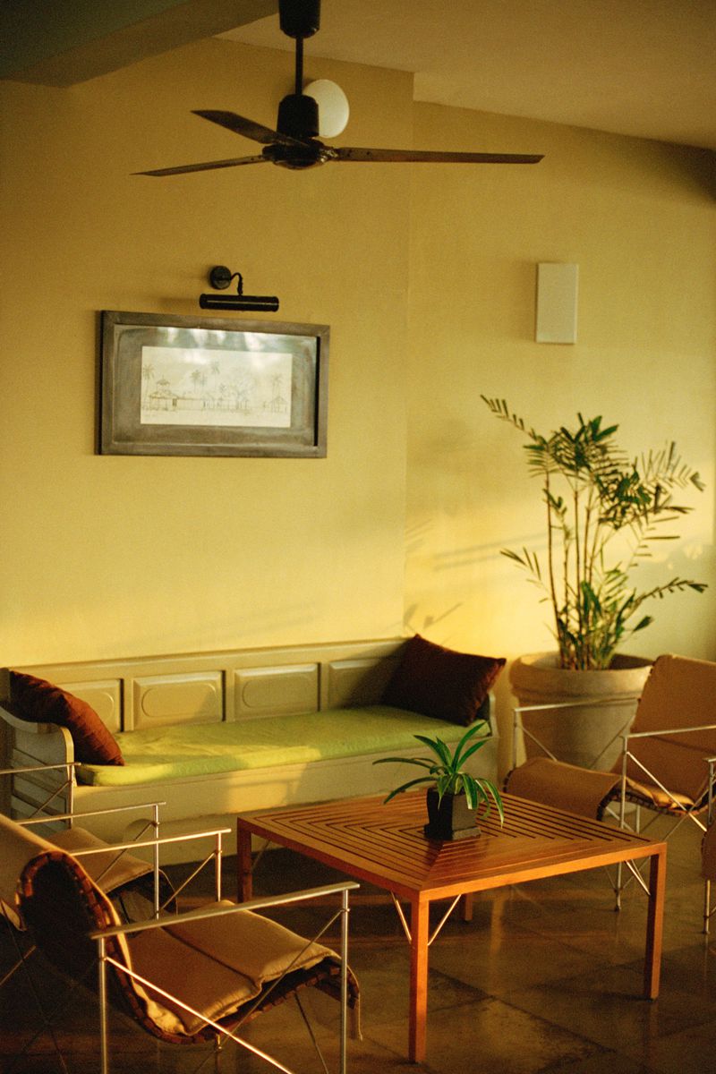 Interior of a room featuring a light green couch with pillows, a wood table, chairs, plants, a framed picture, and a ceiling fan.
