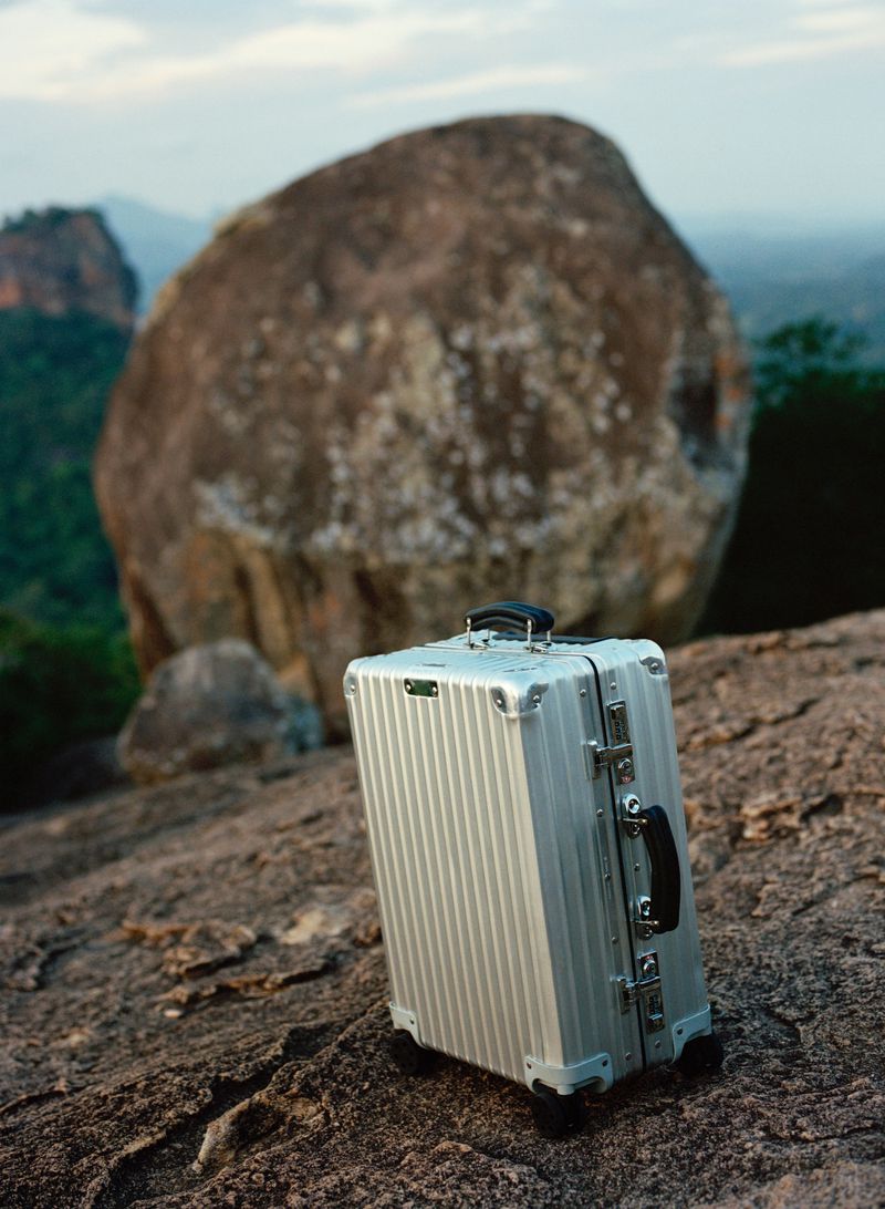 Silver suitcase on a rocky surface, with a large boulder and trees in the soft-focus background. Travel and adventure concept.