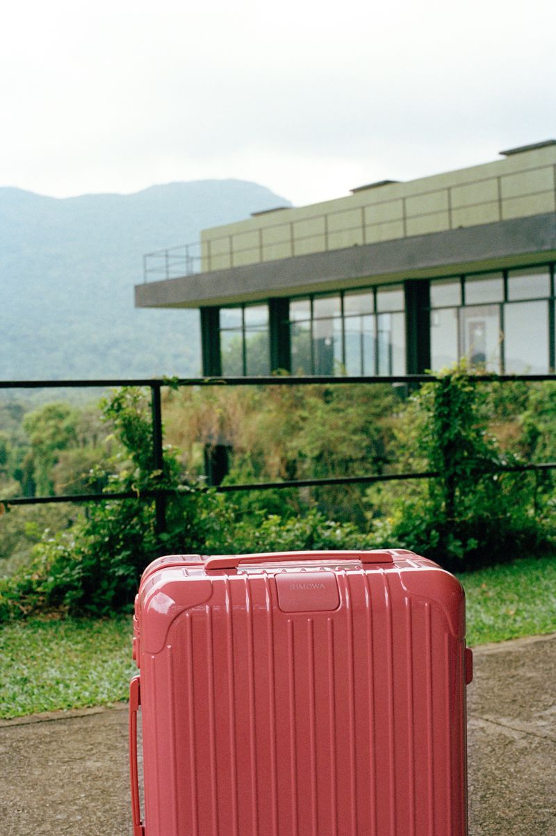 Close-up of a pink suitcase on a patio with a building, green foliage, and mountains in the background on a cloudy day.