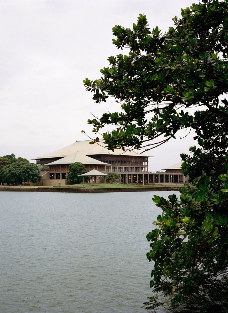 Sri Lanka Parliament building seen from across the water with lush green trees surrounding the architectural landmark on a cloudy day.