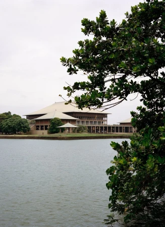 Sri Lanka Parliament building seen from across the water with lush green trees surrounding the architectural landmark on a cloudy day.