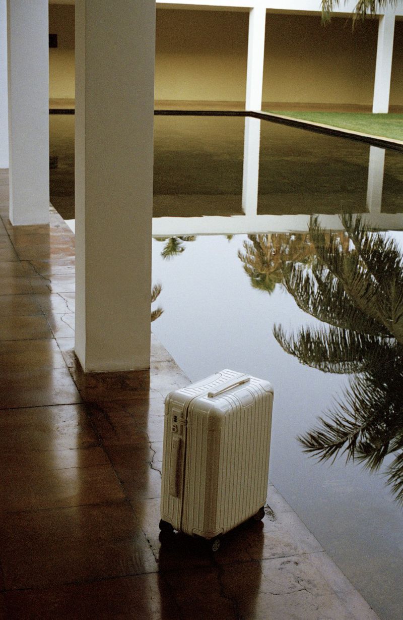 Cream-colored suitcase beside a still pool reflecting white pillars and palm trees, creating a calming travel scene.