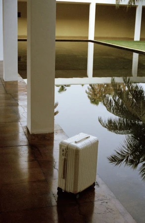 Cream-colored suitcase beside a still pool reflecting white pillars and palm trees, creating a calming travel scene.