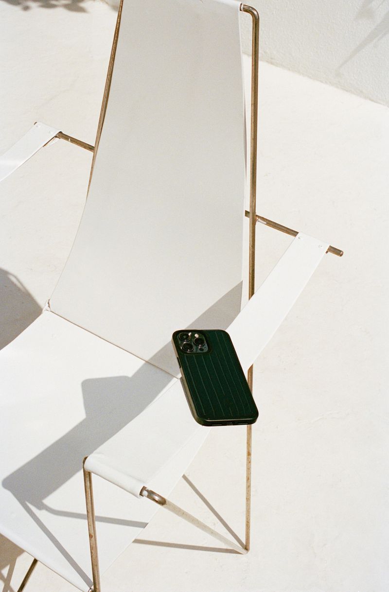 Close-up of a dark green smartphone resting on a bright white sling chair with gold-colored metal frame, captured in full sunlight.