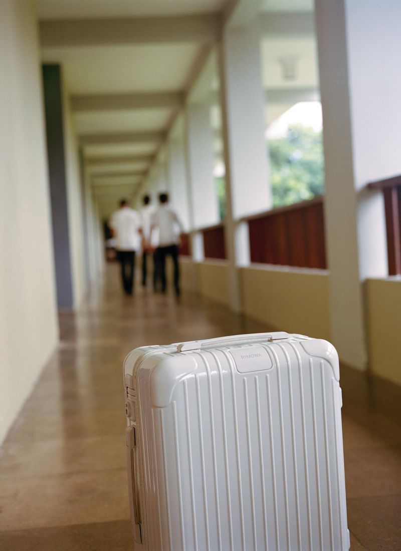 White suitcase in a long hallway with three blurred figures walking in the background. Travel and departure theme in muted colors.