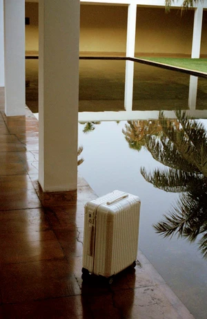 White RIMOWA suitcase standing on a dark tiled floor next to a reflecting pool with architectural pillars.