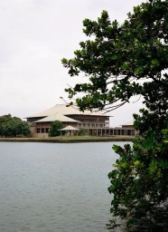 The Sri Lankan Parliament Building across a calm lake, framed by green tree foliage under an overcast sky.