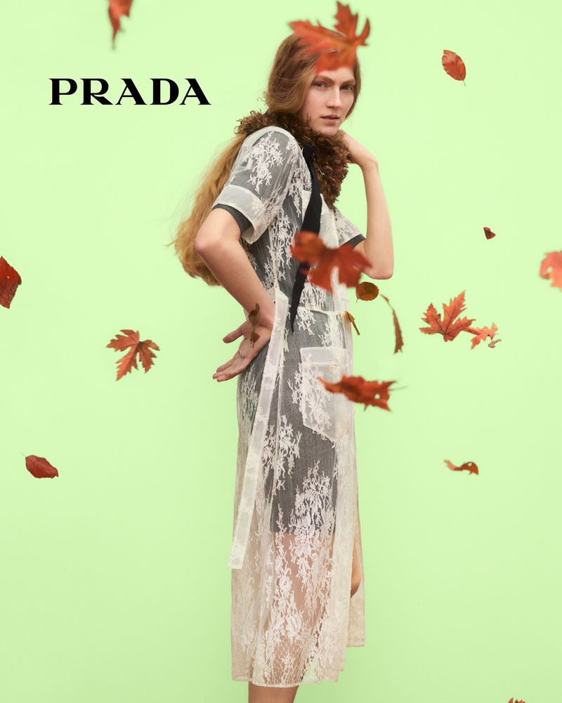 A woman in a lace dress poses in a studio with falling leaves and the Prada logo, creating a fashion-forward and seasonal visual.