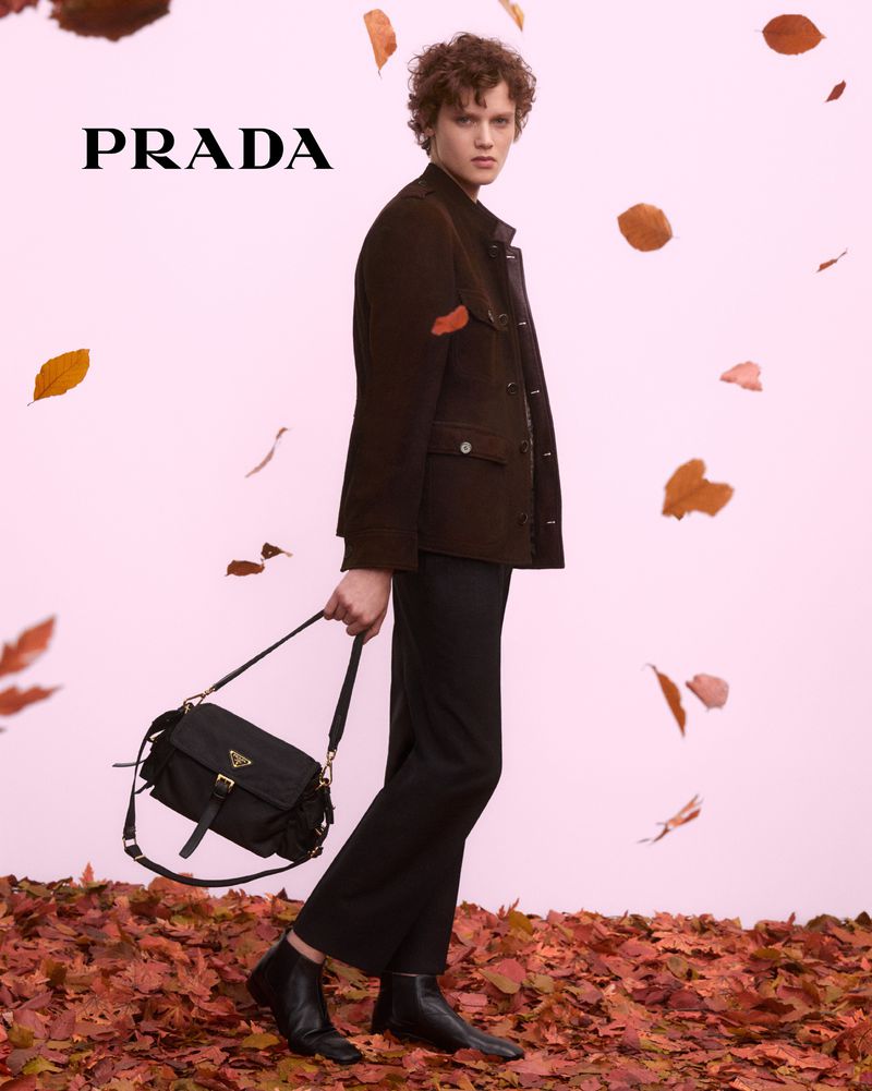 A young adult model stands in a studio surrounded by brown leaves, presenting a black Prada purse in a fashion-style editorial shot.